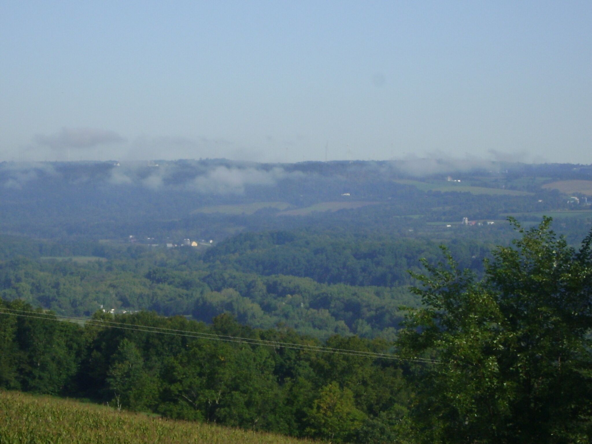 A panoramic view of a hilly landscape with dense forests, a few scattered buildings, and a clear sky.