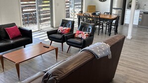 Uma sala de estar moderna com um sofá de couro marrom, duas poltronas de couro preto e uma mesa de centro de madeira. Uma área de jantar com mesa e cadeiras de madeira é visível ao fundo.