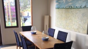 Un comedor con una mesa de madera, seis sillas azules, dos ventanas y un cuadro de paisaje en la pared.