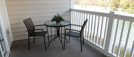 A covered patio with a round table and two chairs, a potted plant, and a view of trees and water.
