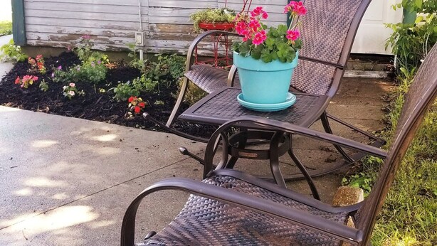 Eine Terrasse mit Metallstühlen und einem Tisch, ein Blumentopf mit Blumen und eine weiße Tür mit grünen Verzierungen.