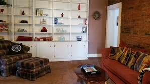 Ein Wohnzimmer mit einem karierten Sofa, einem roten Sitzsofa, einem Couchtisch und einer weißen Regaleinheit mit diversen Gegenständen.