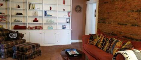 A living room with a plaid sofa, a red couch, a coffee table, and a white shelving unit with various items.