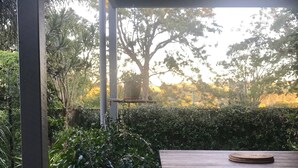 Una terraza de madera con un banco y una mesa, rodeada de plantas en macetas y un gran árbol al fondo.