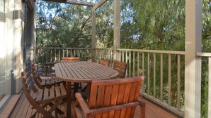 Une terrasse extérieure couverte, avec une table et des chaises en bois, entourée d’une balustrade, et des arbres en arrière-plan.