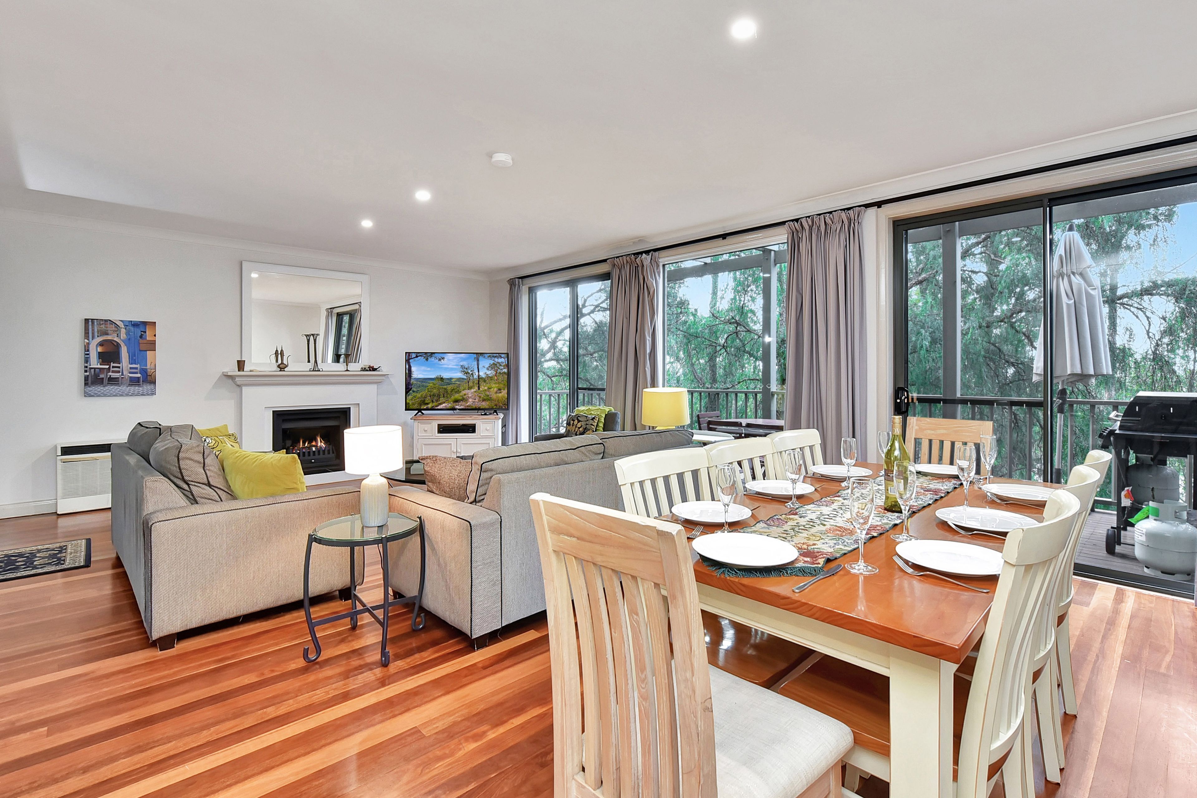 A modern living room with a fireplace, a dining area with a table set for six, and large windows offering a view of the outdoors.