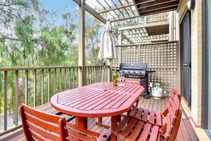 A covered outdoor patio with a red table and benches, a grill, and a bottle of wine.