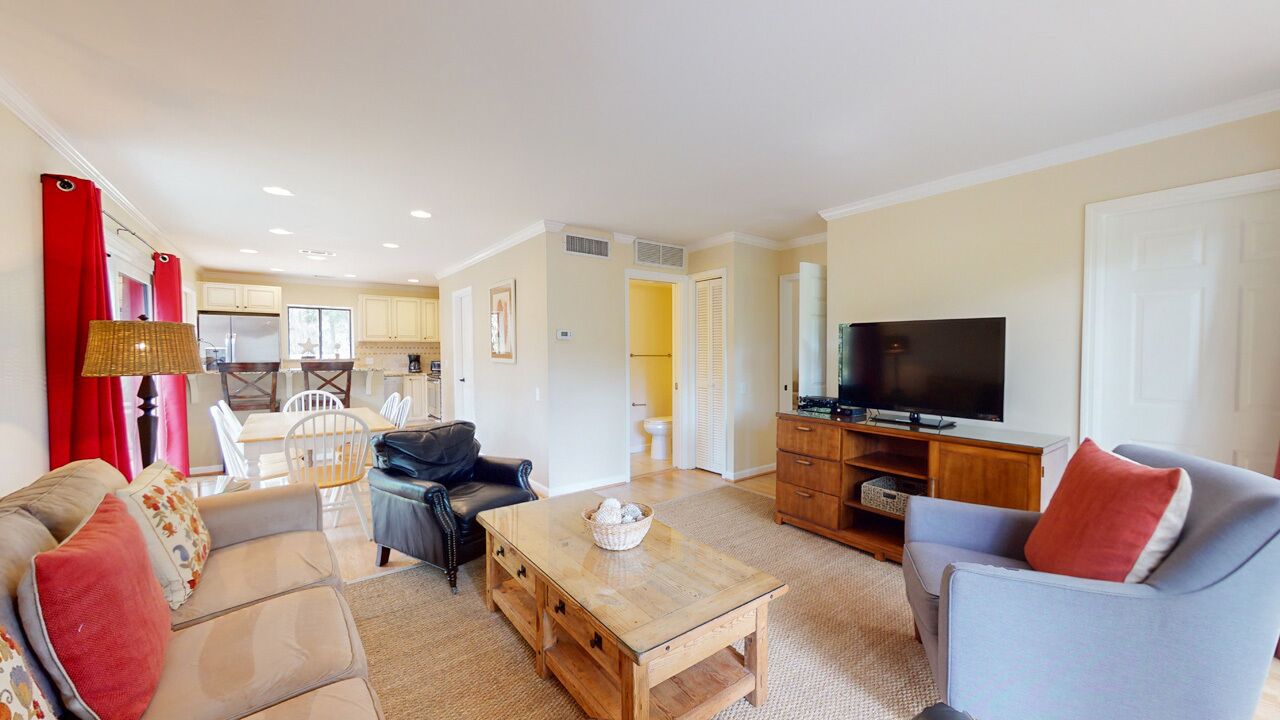 Una sala moderna con sofá, sillón, mesa de centro y televisor. Se observa un área de cocina al fondo.
