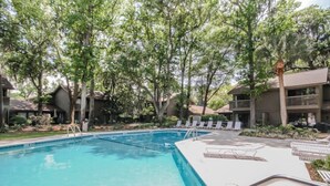 A swimming pool with lounge chairs and a deck area, surrounded by trees and buildings. - Very peaceful and tranquil setting. (Hilton Head)
