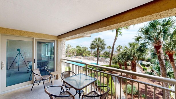 A balcony with a glass door, wicker chairs, a glass table, and a view of a pool and palm trees.