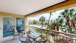 A balcony with a glass door, wicker chairs, a glass table, and a view of a pool and palm trees. - Oceanfront condo, Two Bedroom, Second Floor (Hilton Head)