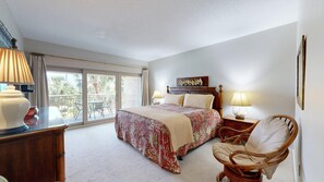 A bedroom with a bed, two bedside lamps, a chair, a dresser, and a balcony with chairs. - Oceanfront condo, Two Bedroom, Second Floor (Hilton Head)