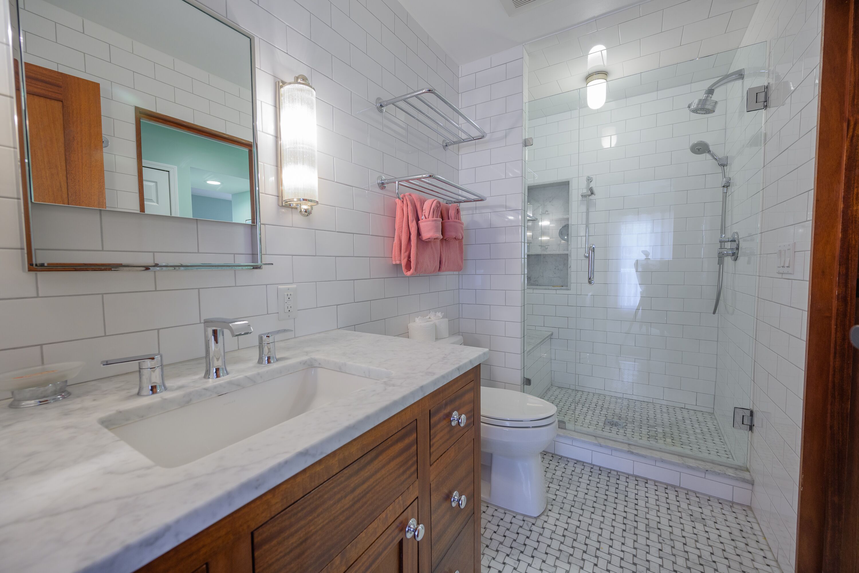 A bathroom with a white tiled wall, a wooden vanity with a sink, a mirror, a towel rack with pink towels, a toilet, and a glass-enclosed shower area.