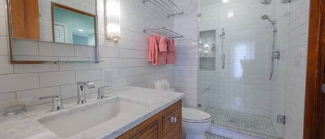A bathroom with a white tiled wall, a wooden vanity with a sink, a mirror, a towel rack with pink towels, a toilet, and a glass-enclosed shower area.