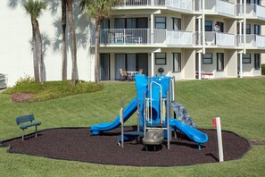 Um parquinho com um escorrega azul e um banco, cercado por um jardim com palmeiras e um edifício com varandas ao fundo.