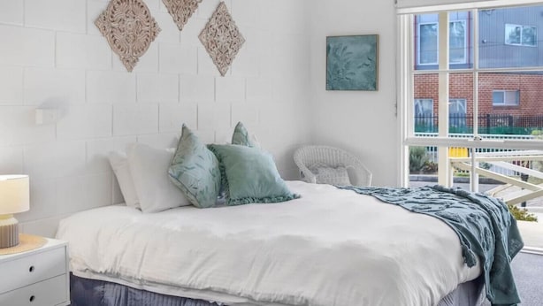 Une chambre à coucher comprenant un lit, des oreillers, une table de chevet, une chaise et une fenêtre donnant sur des immeubles.