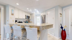 A kitchen with white cabinets, a microwave, a stainless steel refrigerator, and a granite countertop. There are wicker chairs around the island. - Fifth Floor Condo With Great Views of the Atlantic Ocean! (Hilton Head)
