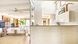 A spacious living area with a wooden floor, a sofa set, a dining table, and a kitchen area with white cabinets and a large window. - AZURE - BEACH HOUSE ON THE BEACH (Marcus Beach)