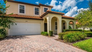A two-story house with a beige facade, a red-tiled roof, and a white garage door. There are two windows on the upper floor and a covered entryway with arches. - Executive 5 Bed 4.5 Bath Vacation Home W/ Pool, Spa Near Disney  (Kissimmee)