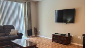 A living room with a wooden floor, a dark sofa, a coffee table, a TV mounted on the wall, and a ceiling fan. - Executive 5 Bed 4.5 Bath Vacation Home W/ Pool, Spa Near Disney  (Kissimmee)