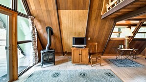A cozy attic room with a wooden sloped ceiling, a black wood-burning stove, a television on a wooden cabinet, and a small table with a lamp. - Serene Washington Escape w/ Deck & Mountain Views! (Packwood)