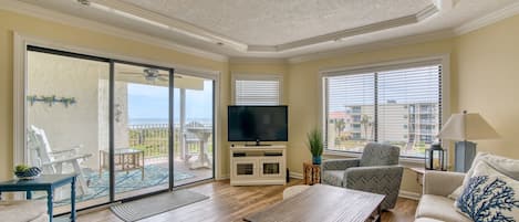 A spacious living room with a large sliding glass door leading to a balcony, a flat-screen TV, and a comfortable seating area with a sofa and chairs.