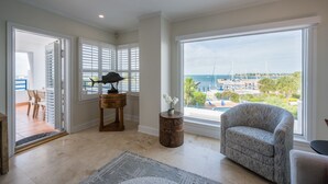A spacious living room with a large window offering a marina view, a comfortable armchair, a small side table with a vase of flowers, and a wooden side table with a lamp.
