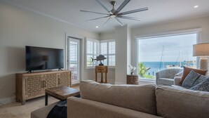 A living room with a large flat-screen TV, a ceiling fan, a sofa, a wooden cabinet, and a view of the ocean through large windows.