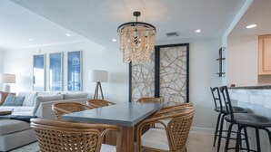 A modern dining area with a wooden table and wicker chairs, a chandelier, and a view of the city skyline through large windows.