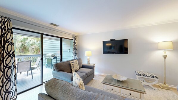 Een woonkamer met een grijze bank, een glazen salontafel, een flatscreen-tv en een lamp. Er is een schuifdeur die leidt naar een buitenpatio met een tafel en stoelen.