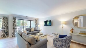 Un salon avec un canapé gris, un fauteuil à motifs, une table basse, un téléviseur fixé au mur, une lampe sur pied et un miroir au-dessus d’une console.