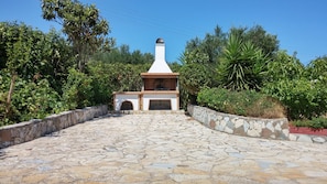 Un chemin de pierres mène à un espace barbecue extérieur aux couleurs blanc et marron, entouré d’une végétation luxuriante.