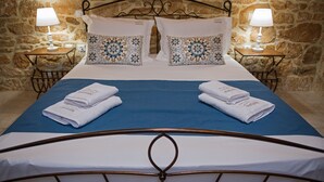 Een netjes opgemaakt bed met twee kussens, een blauwe dekbedovertrek en twee gevouwen handdoeken met de naam van het hotel erop. Twee nachtkastjes met lampen en een stenen muur op de achtergrond.