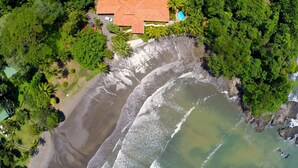 An aerial view of a coastal property with a beach, dense greenery, and a building with a red roof. - SOUTH BEACH PARADISE! DIRECT OCEANFRONT WITH COMPLIMENTARY DAILY CHEF BREAKFAST! (Jaco Beach)