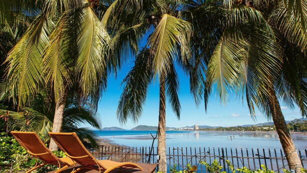 A wooden deck with two wicker lounge chairs facing a calm body of water, surrounded by palm trees and a clear blue sky. - SOUTH BEACH PARADISE! DIRECT OCEANFRONT WITH COMPLIMENTARY DAILY CHEF BREAKFAST! (Jaco Beach)