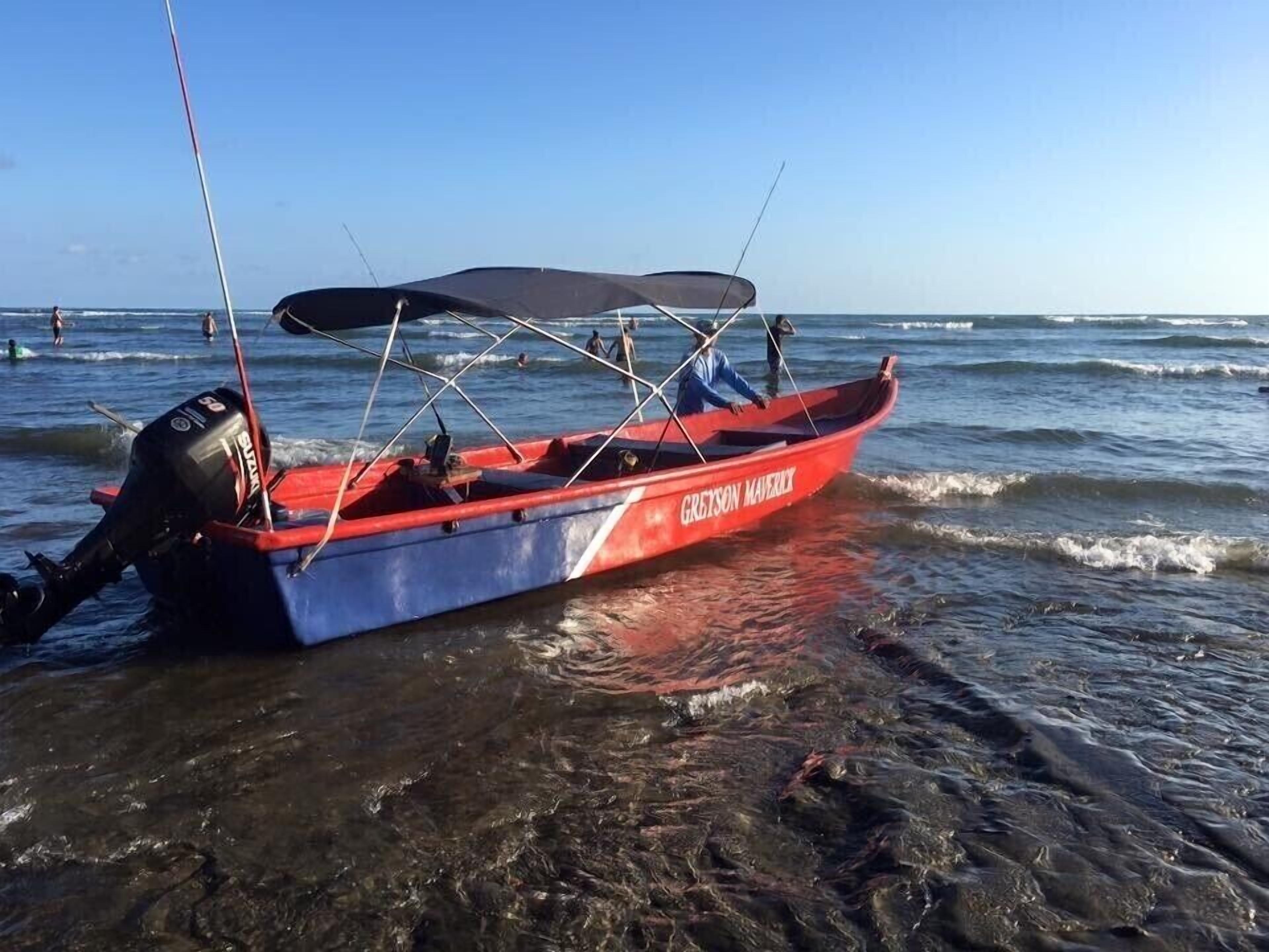 Una piccola imbarcazione rossa e blu con una tenda, chiamata "GREYSON MAJERY", ormeggiata su una spiaggia con acque limpide e alcune persone sullo sfondo.
