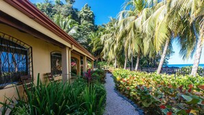 A pathway leading to a building with a tiled roof, surrounded by lush greenery and palm trees. - SOUTH BEACH PARADISE! DIRECT OCEANFRONT WITH COMPLIMENTARY DAILY CHEF BREAKFAST! (Jaco Beach)