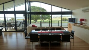 A dining area with a large table and chairs, a sofa, and a view of a road and greenery through large windows.