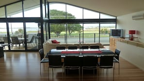 A dining area with a large table and chairs, a sofa, and a view of a road and greenery through large windows. - GoolwaGettaway Luxury Home by river- sleeps 14 in 10 beds (Goolwa South)