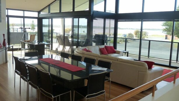 A spacious living area with a glass wall, a dining table with chairs, and a sofa with red cushions.