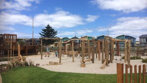 A playground with wooden structures and a sandy area, surrounded by buildings with modern architecture.