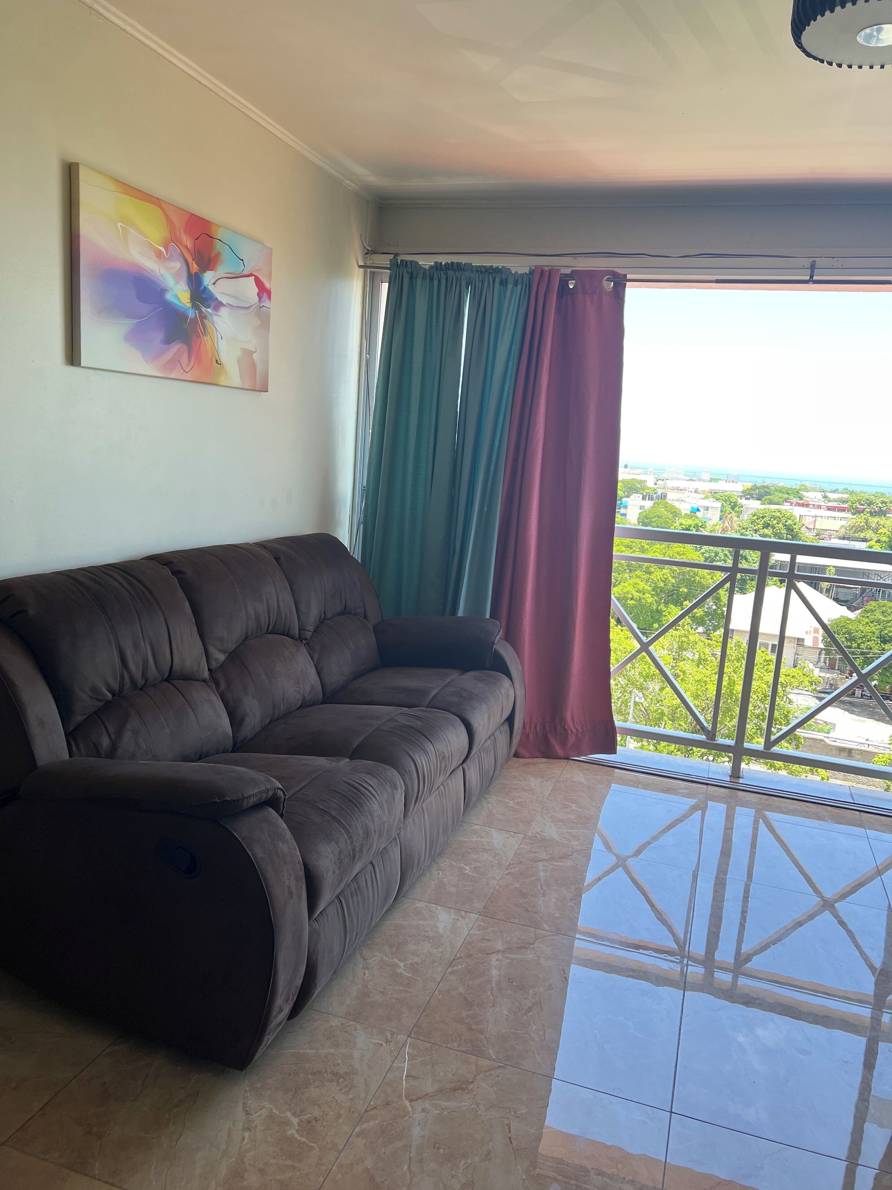 A living room with a brown sofa, a colorful abstract painting, and a view of a cityscape through a balcony with green and purple curtains.
