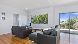 Una sala moderna con dos sofás grises, una mesa de centro de madera y un televisor de pantalla plana. Amplias ventanas ofrecen vistas a árboles y un balcón.