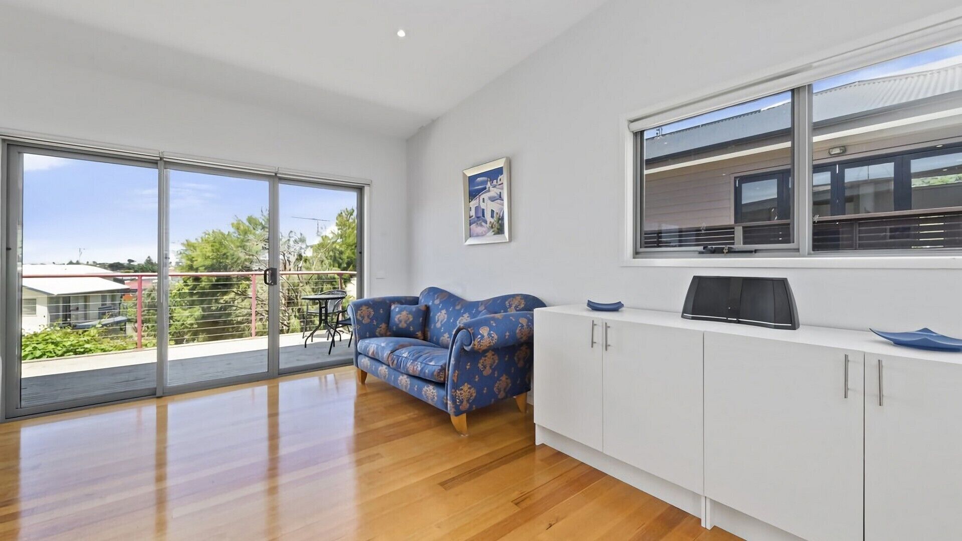 A spacious living area with a large sliding glass door leading to a balcony, a blue patterned sofa, a white cabinet, and a framed picture on the wall.