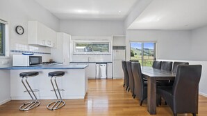 Una cocina moderna con armarios blancos, microondas y pileta. Hay un comedor con mesa y sillas, y vistas al exterior a través de amplios ventanales.