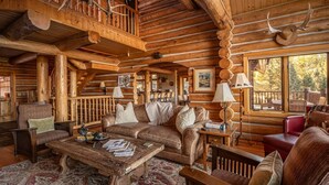 A rustic log cabin interior with a wooden staircase, leather sofas, a coffee table, and a large window offering a view of the outdoors. - Enchanting Private Mountain Sanctuary! Spectacular Views (Big Sky)