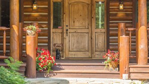 A wooden cabin with a large front door, two windows, and a porch with wooden posts and flower pots. - Enchanting Private Mountain Sanctuary! Spectacular Views (Big Sky)