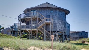 A wooden, multi-story house with a balcony and stairs, surrounded by grass and other houses. - Point Break Cottage- Steps to Beach- Community Pool & Spa (Buxton)