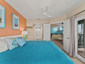 A bedroom with a bed, a ceiling fan, a bathroom door, and a view of a balcony.