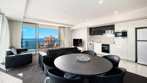 A modern living room with a round table, black chairs, and a sofa. There is a kitchen area with white cabinets and a refrigerator. A large window offers a view of the city and a building across the street. - H Residences Level 10, Ocean, Free Wi-Fi, Parking, Foxtel (Gold Coast)